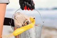 Person wearing red and black gloves holding a Boardwalk Boutique Hotel branded metal bucket filled with beach cleanup items on an Aruba beach