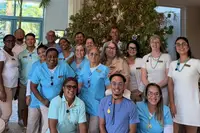Boardwalk Boutique Hotel Aruba team celebrating in the lobby in front of the decorated Christmas tree, waving and smiling under tropical chandeliers.