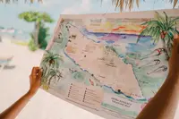 Hands holding an illustrated treasure map on the beach at Boardwalk Boutique Hotel Aruba, with sea and palm trees in the background.