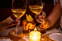 Two guests clink glasses of white wine at a candlelit table at The Coco Cafe, Boardwalk Boutique Hotel Aruba, with small dishes on the table.