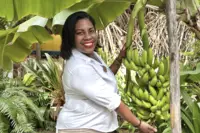 Smiling team member in the tropical garden holding a bunch of green bananas at Boardwalk Boutique Hotel Aruba.