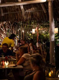 Lively outdoor restaurant at night under a thatched roof with string lights. 