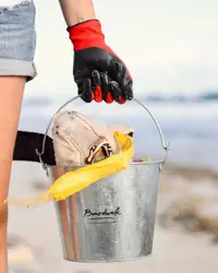 Person wearing red and black gloves holding a Boardwalk Boutique Hotel branded metal bucket filled with beach cleanup items on an Aruba beach