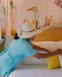 Hotel staff member in a blue uniform and sun hat arranging pillows on a bed, with a colorful bird mural in the background